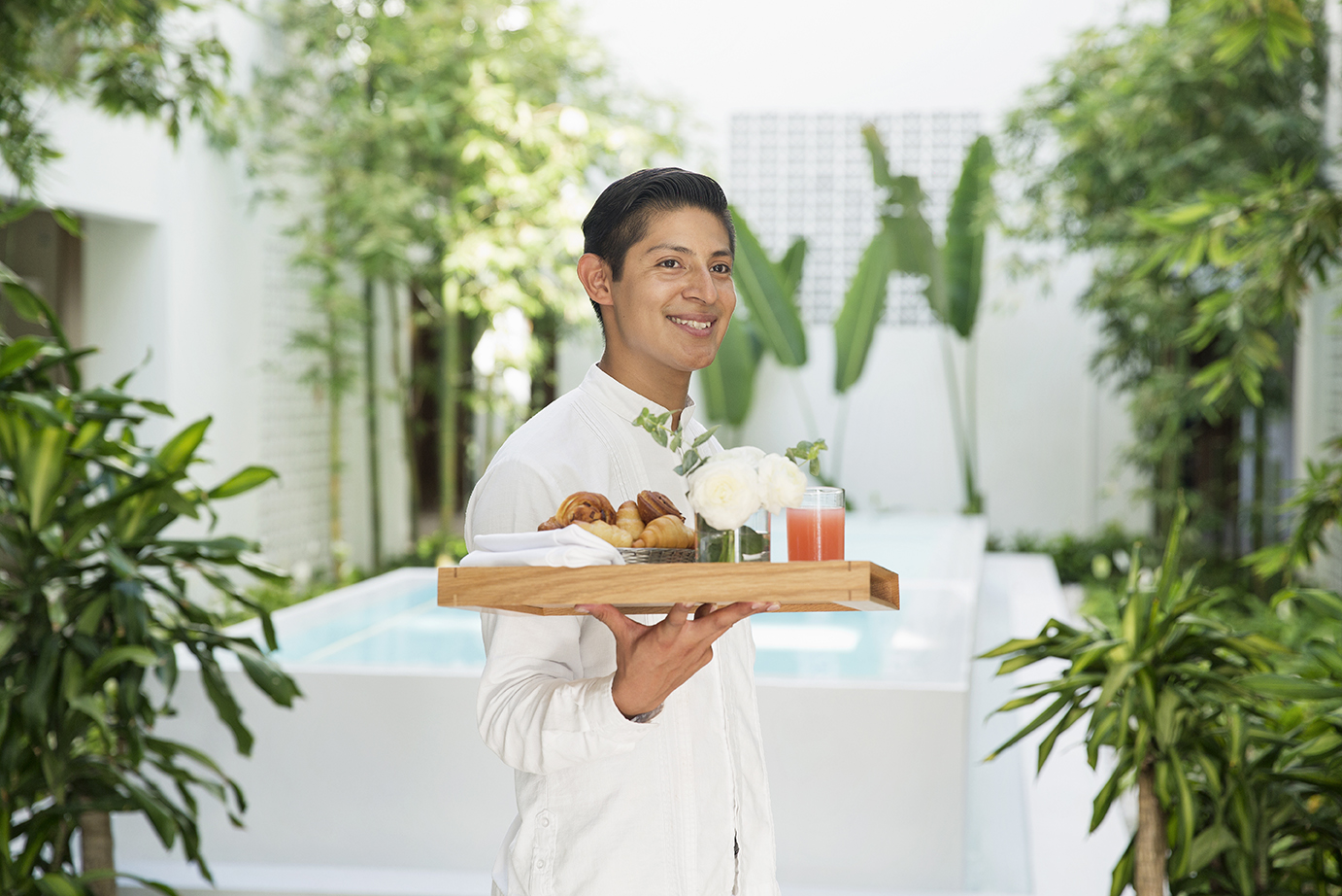 Server holding wooden tray with pastries, juice, and flowers in lush garden courtyard.